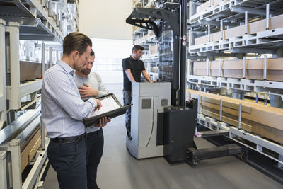 Two men with documents and worker with forklift in factory warehouse