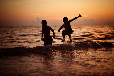 Silhouette boys playing in sea against sky during sunset