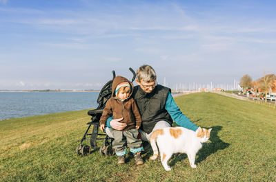 Grandfather and granddaughter with cat on grassy field by lake against sky