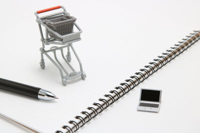 Close-up of toy laptop by shopping cart and spiral notebook against white background