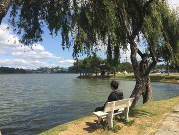 Man sitting on bench by lake against sky