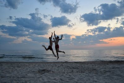 Men jumping at beach against sky during sunset