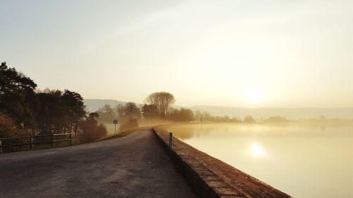 Empty road by calm lake against sky during sunrise