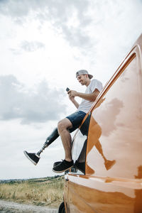 Young man with leg prosthesis sitting on roof of camper van using cell phone