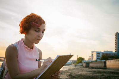 Mid adult woman writing while sitting at riverbank against sky during sunset