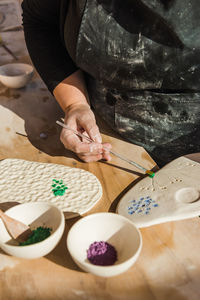 Crop anonymous female artisan in dirty black apron using paintbrush for painting ceramic palette on table near bowls with pigment in bright workshop