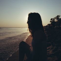 Side view of young woman sitting at beach during sunset