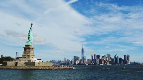 Statue of liberty by sea with city at waterfront against cloudy sky