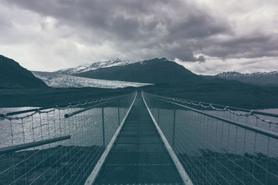 Empty footbridge leading towards mountains against sky