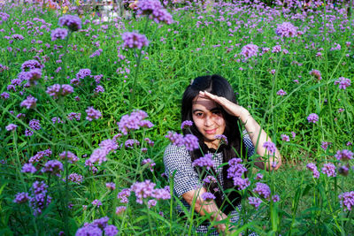 Portrait of smiling woman shielding eyes while sitting amidst purple flowers on field