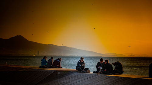 Silhouette people sitting on sea against orange sky