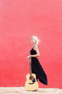 Vertical photo of a young beautiful blond woman in a black dress standing leaning on a guitar
