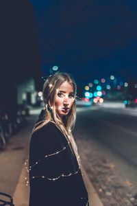 Side view portrait of young woman with illuminated string lights standing on street at night