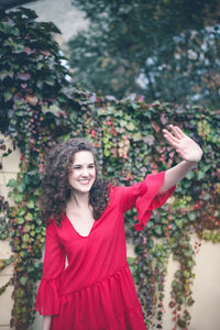 Smiling young woman in red dress walking outdoors, waving her hand. curly hair brunette, autumn