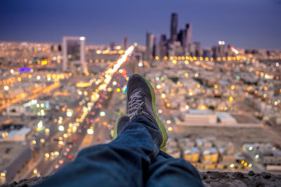 Low section of man sitting on cliff against illuminated cityscape