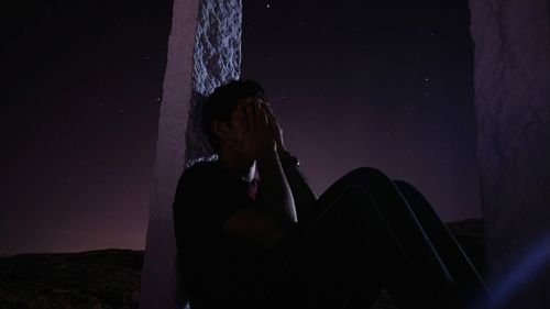 Man with face covered by hands sitting against sky at night