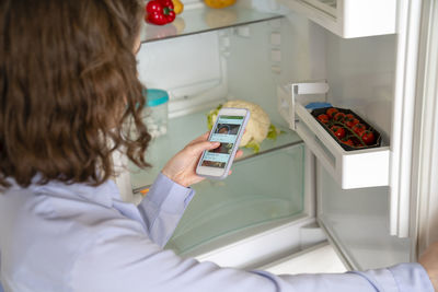 Woman doing online shopping of groceries through smart phone by refrigerator