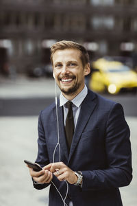 Portrait of smiling entrepreneur using smart phone through in-ear headphones while standing outdoors