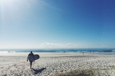 Rear view of man with surfboard walking on sand at beach against sky during summer