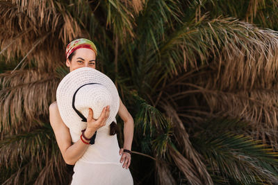 Portrait of woman holding hat against tree