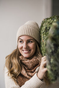 Smiling blond woman carrying christmas tree on shoulder at home