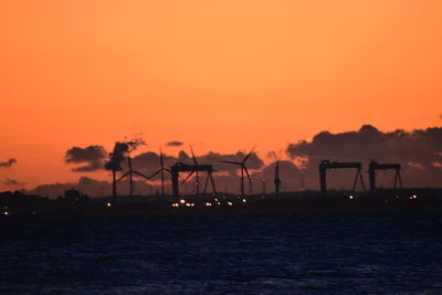 Silhouette cranes by sea against orange sky