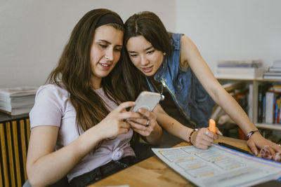 Young female friends sharing smart phone while studying together at home