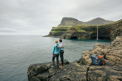 Rear view of people standing on rock by sea against sky