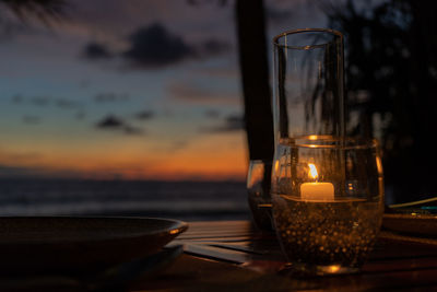 Close-up of burning candle in glass container on table against sky during sunset