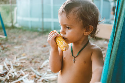 Cute shirtless baby boy eating corn while standing outdoors