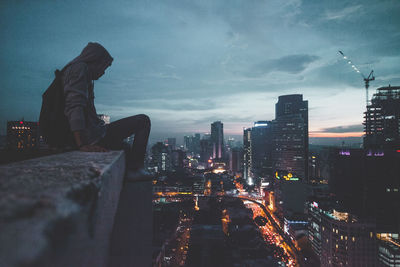 Full length of man sitting on building terrace by illuminated cityscape against sky during sunset