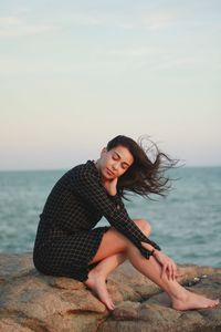 Young woman sitting on shore at beach against sky