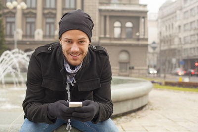 Man using mobile phone while sitting against fountain