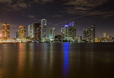 Illuminated buildings by river against sky at night