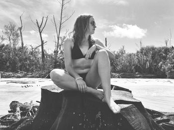 Young woman wearing bikini while sitting on tree stump at beach against sky