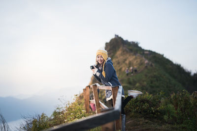 Portrait of smiling young woman with camera standing on mountain against clear sky