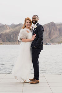 Side view portrait of happy newlywed couple standing on pier over lake