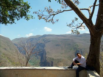 Portrait of man sitting on retaining wall with trees and mountains in background