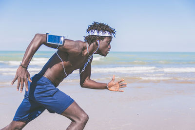 Side view of man listening music while running on shore at beach against sky