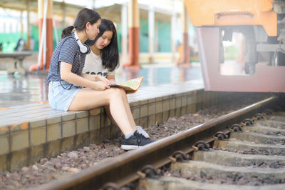Two teenage female travelers look at maps on a train platform.