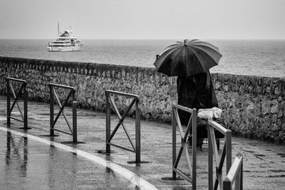 Woman in rain with umbrella and shopping cart with yacht on the sea in background