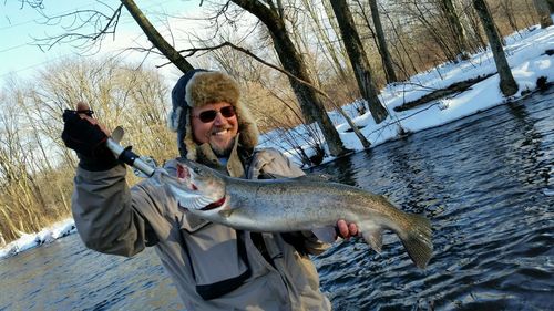 Happy mature man holding steelhead trout at river against bare trees during winter