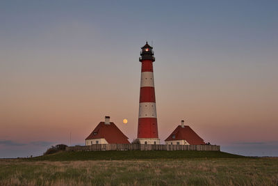 Lighthouse on field by building against sky during sunset