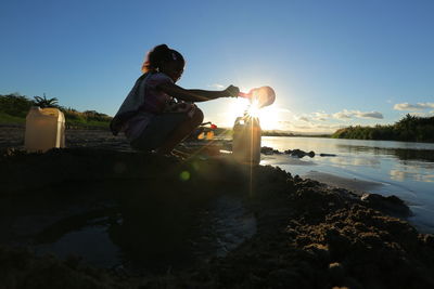 Side view of woman filling water in can while crouching on lakeshore