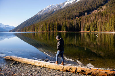 Side view of young woman walking on log at lakeshore against mountain