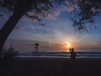 Silhouette of man enjoying the sunrise at the beach and using smartphone on tripod to take pictures.