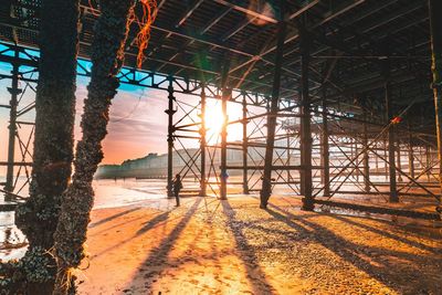 Silhouette woman standing below pier at beach during sunset