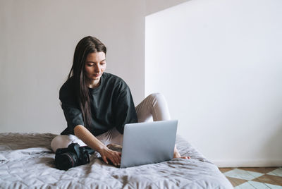 Young woman photographer with digital camera freelancer in casual using laptop on bed at home