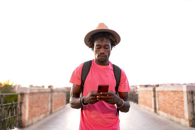 Handsome african american male traveler with backpack standing on bridge and using smartphone