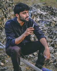 Thoughtful young man smoking cigarette while siting on fallen tree trunk